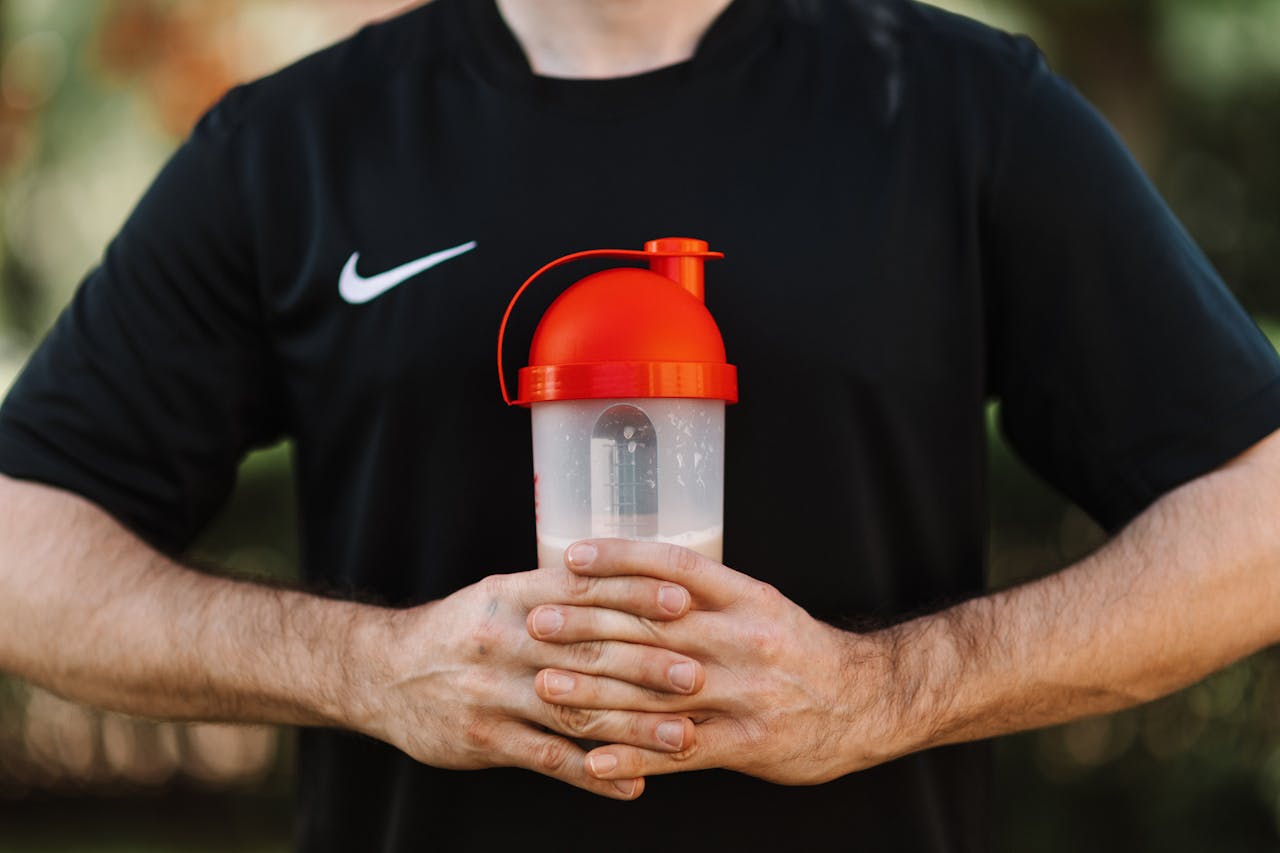 Close-up of a man holding a protein shaker outdoors, wearing a black shirt.