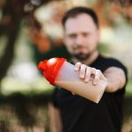 Man presenting a protein shake in a plastic tumbler against a blurred nature background.