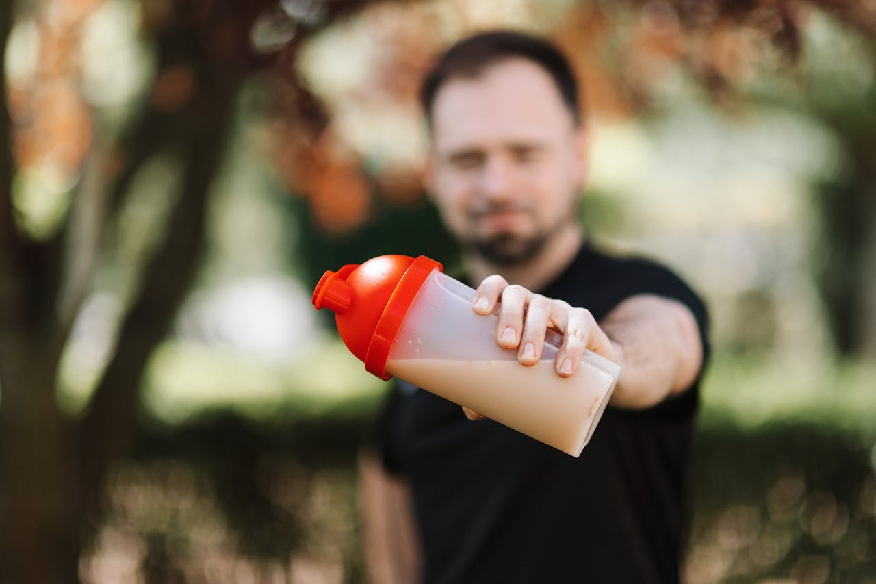 Man presenting a protein shake in a plastic tumbler against a blurred nature background.
