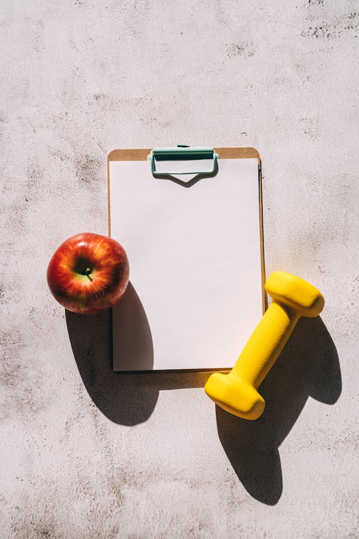 Flat lay of apple, clipboard, and dumbbell representing fitness and nutrition concept.